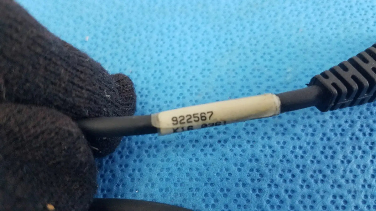 Gloved hand holding black cable with beige label 922567 for 09840-804 barcode scanner pen with rj connector against blue background