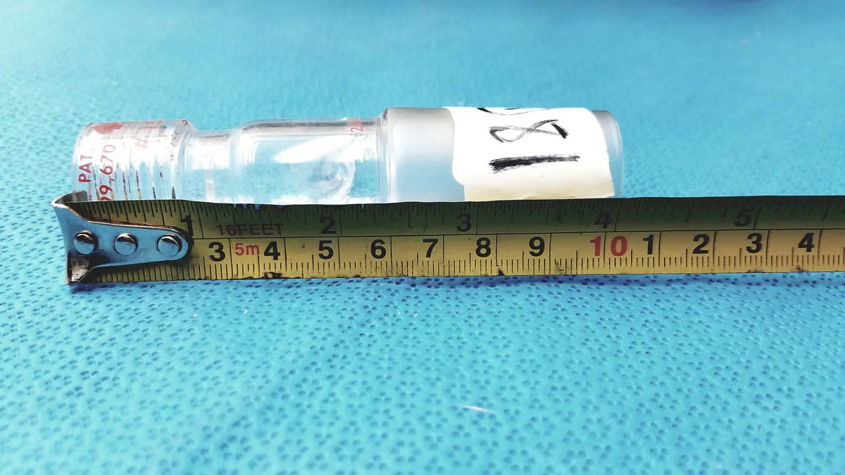 Clear glass ace glass chem-thread inlet adapter with white label and red cap text beside yellow measuring tape in laboratory glassware