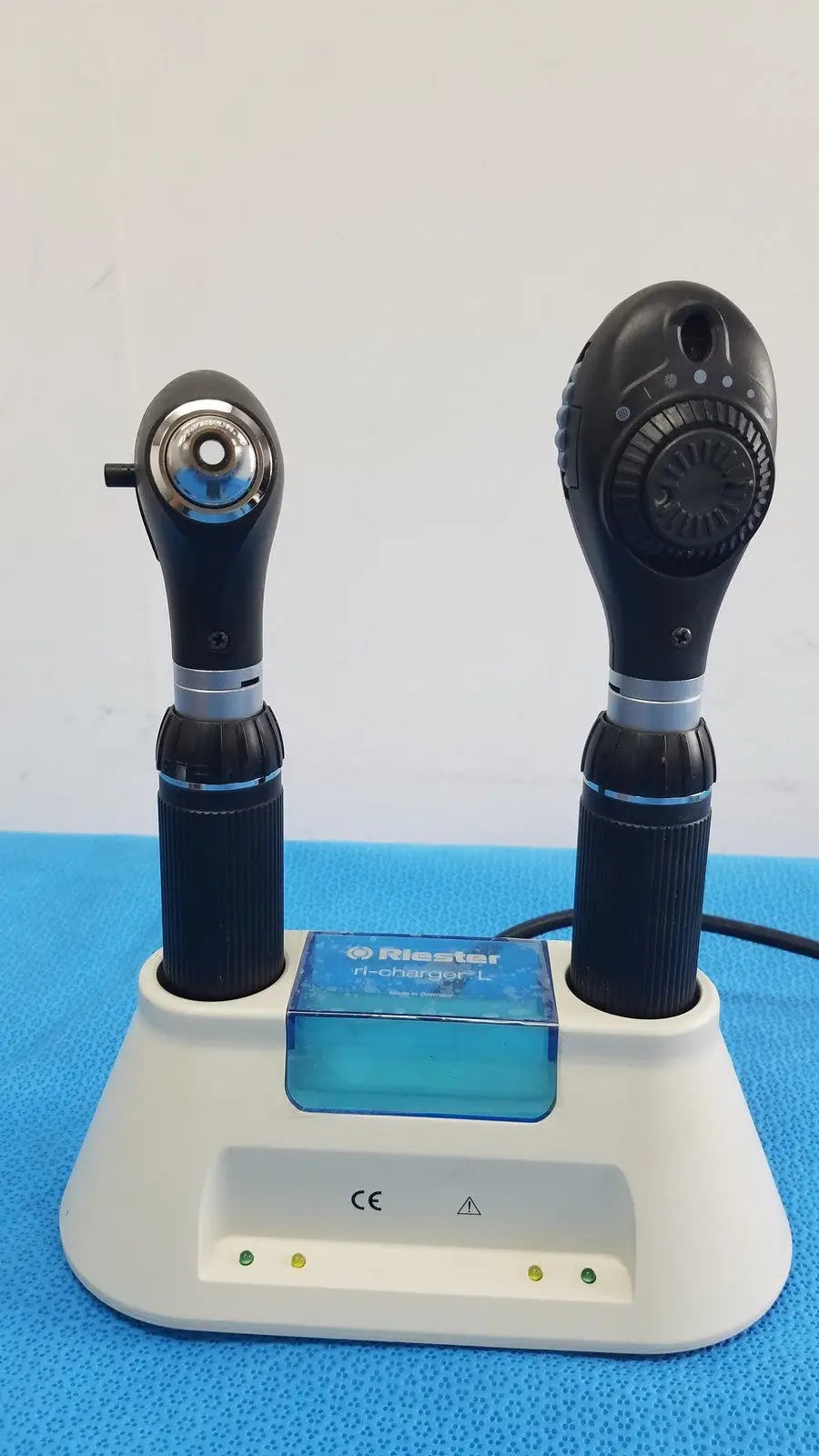 Two black rieker h-checker l otoscopes with textured grips and silver accents sit in a white ri-charger l charging base