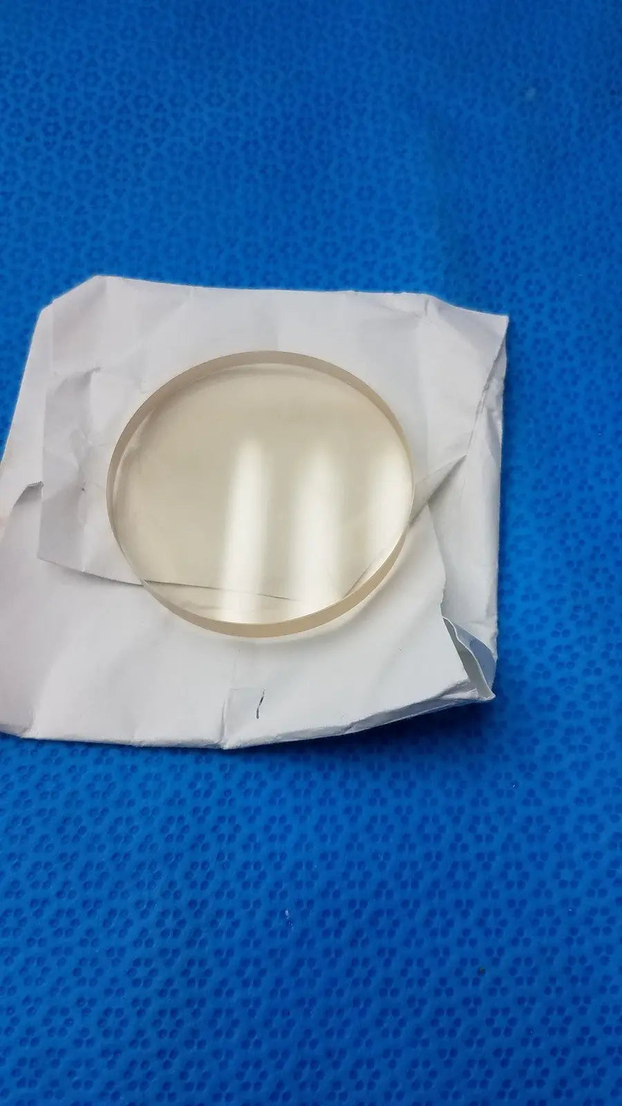 Clear circular glass lens w1147 schott zerodur exp class 2 127x90mm resting on crumpled white paper wrapper