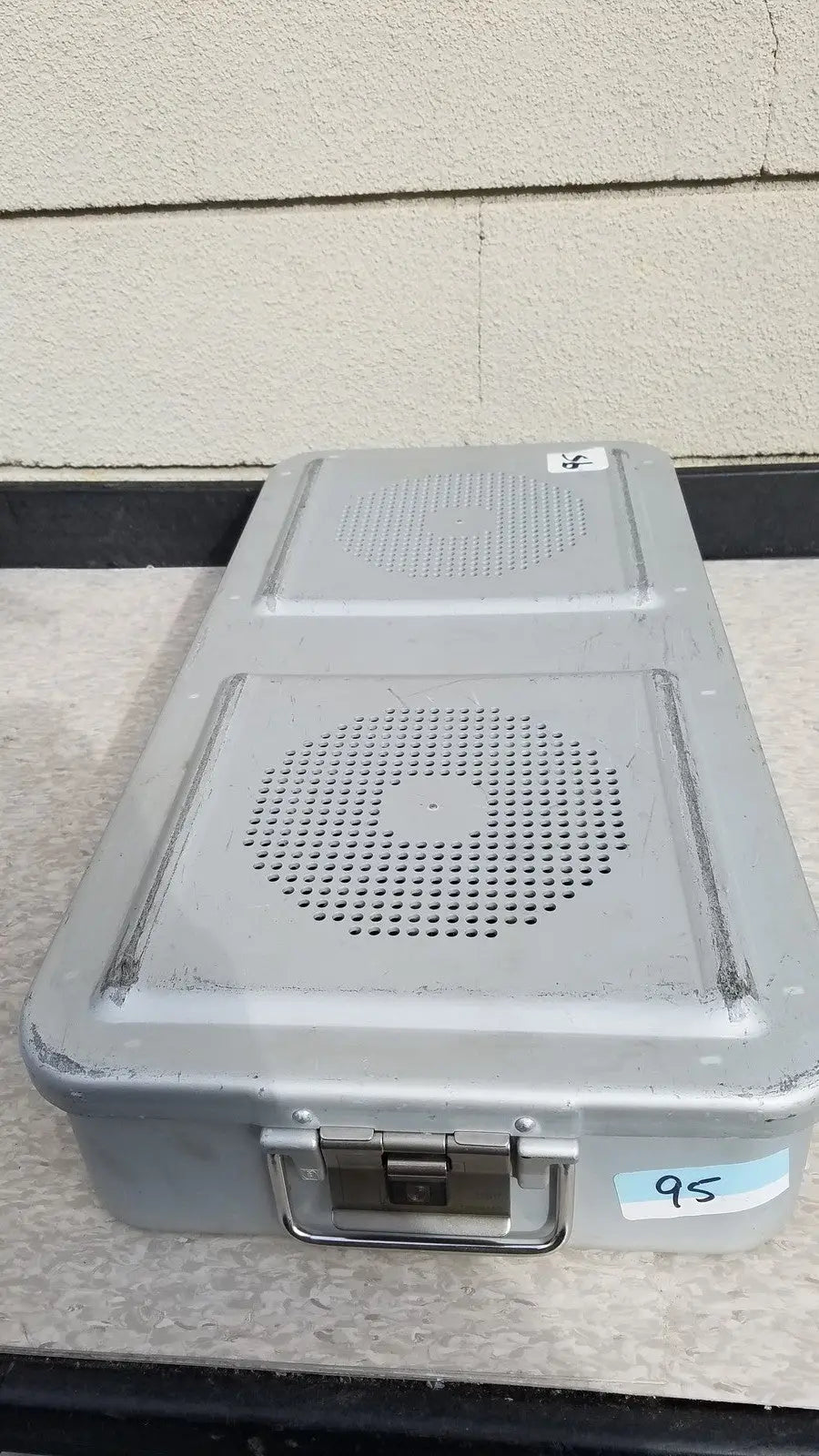 Gray metal aesculap jn821 sterilization case with two circular perforated speaker grilles and handle on top surface