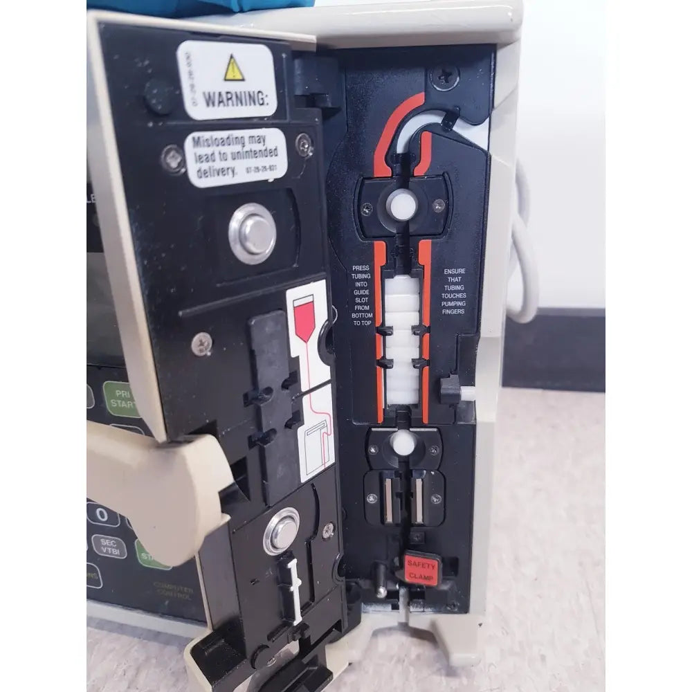Black and beige baxter 2m8063 volumetric infusiion pump with orange tubing guide safety clamp and warning labels