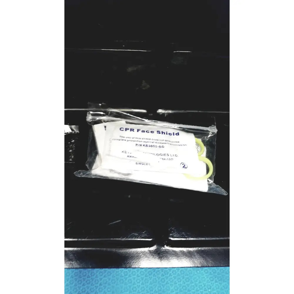 A white cpr face shield with yellow elastic strap in clear packaging for defibrillator storage cabinet wall mount