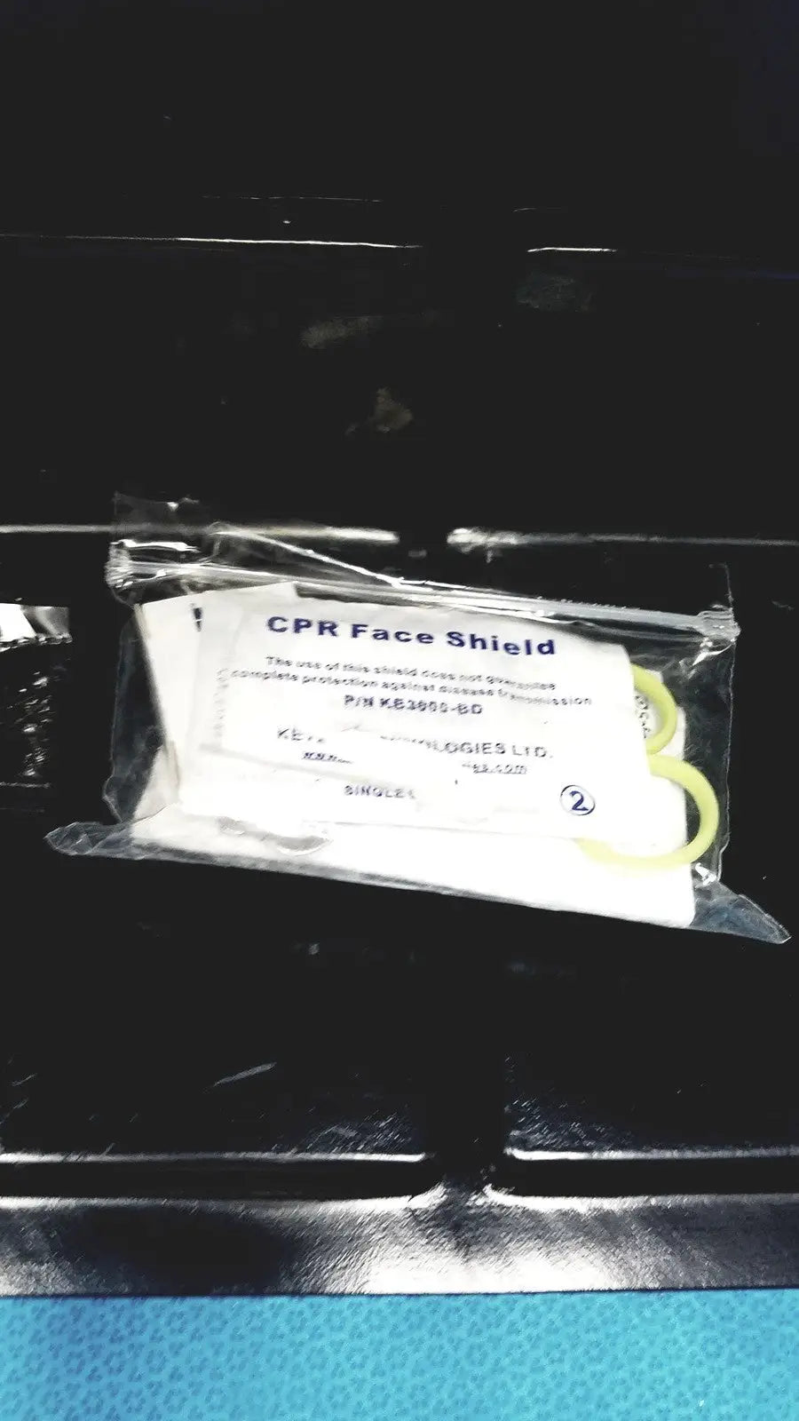 A white cpr face shield with yellow elastic strap in clear packaging for defibrillator storage cabinet wall mount