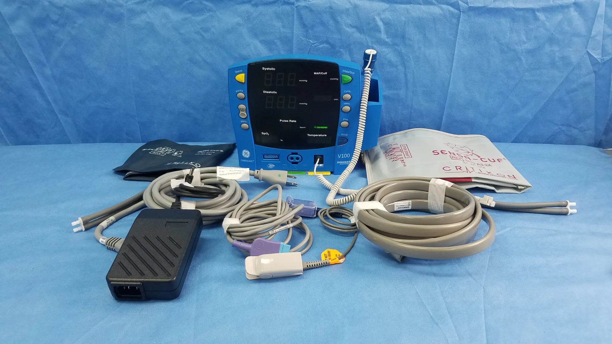 Blue GE Dinamap V100 vital signs patient monitor with SpO2 sensor, nibp cuffs, power adapter, and patient cables on blue surface