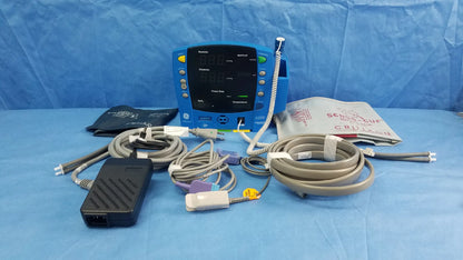 Blue GE Dinamap V100 vital signs patient monitor with SpO2 sensor, nibp cuffs, power adapter, and patient cables on blue surface