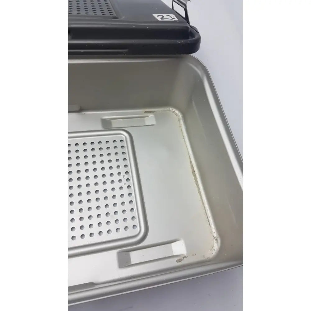 Black plastic lid with white number 21 sticker on silver perforated tray of genesis solid brass sterilization container