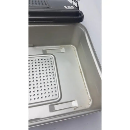 Black plastic lid with white number 21 sticker on silver perforated tray of genesis solid brass sterilization container
