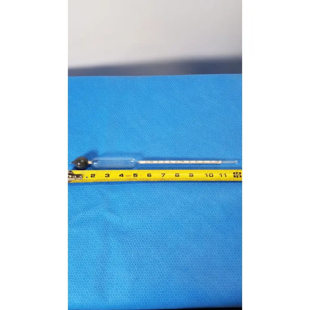 A clear glass specific gravity hydrometer with a black weighted bulb resting on a yellow tape for light liquids temp measurement