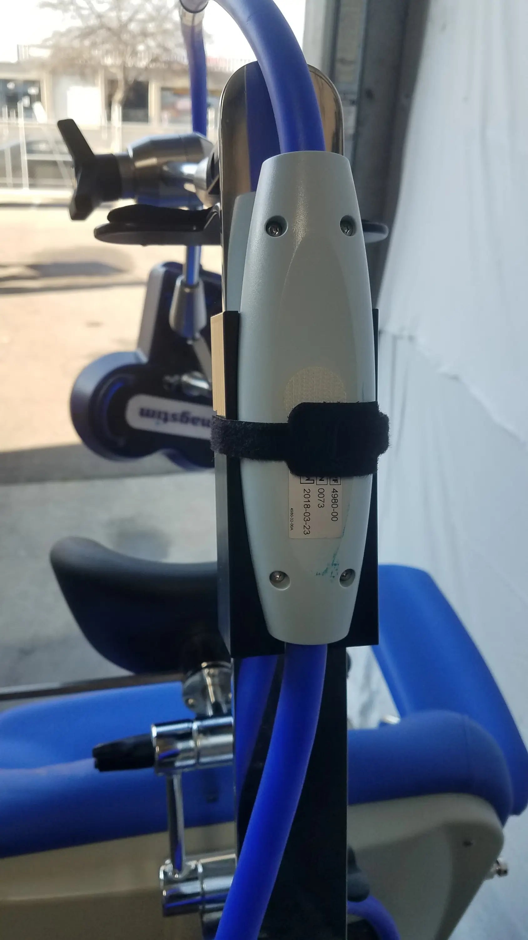 White cylindrical stimulation coil with black strap mounted on blue tubing attached to magstim therapy chair articulating arm