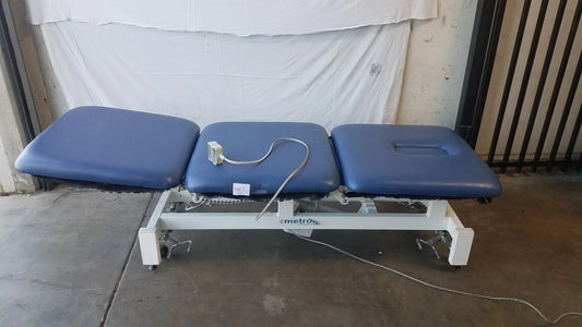 Blue padded three-section metro electric treatment table with white metal frame and wheels for medical use