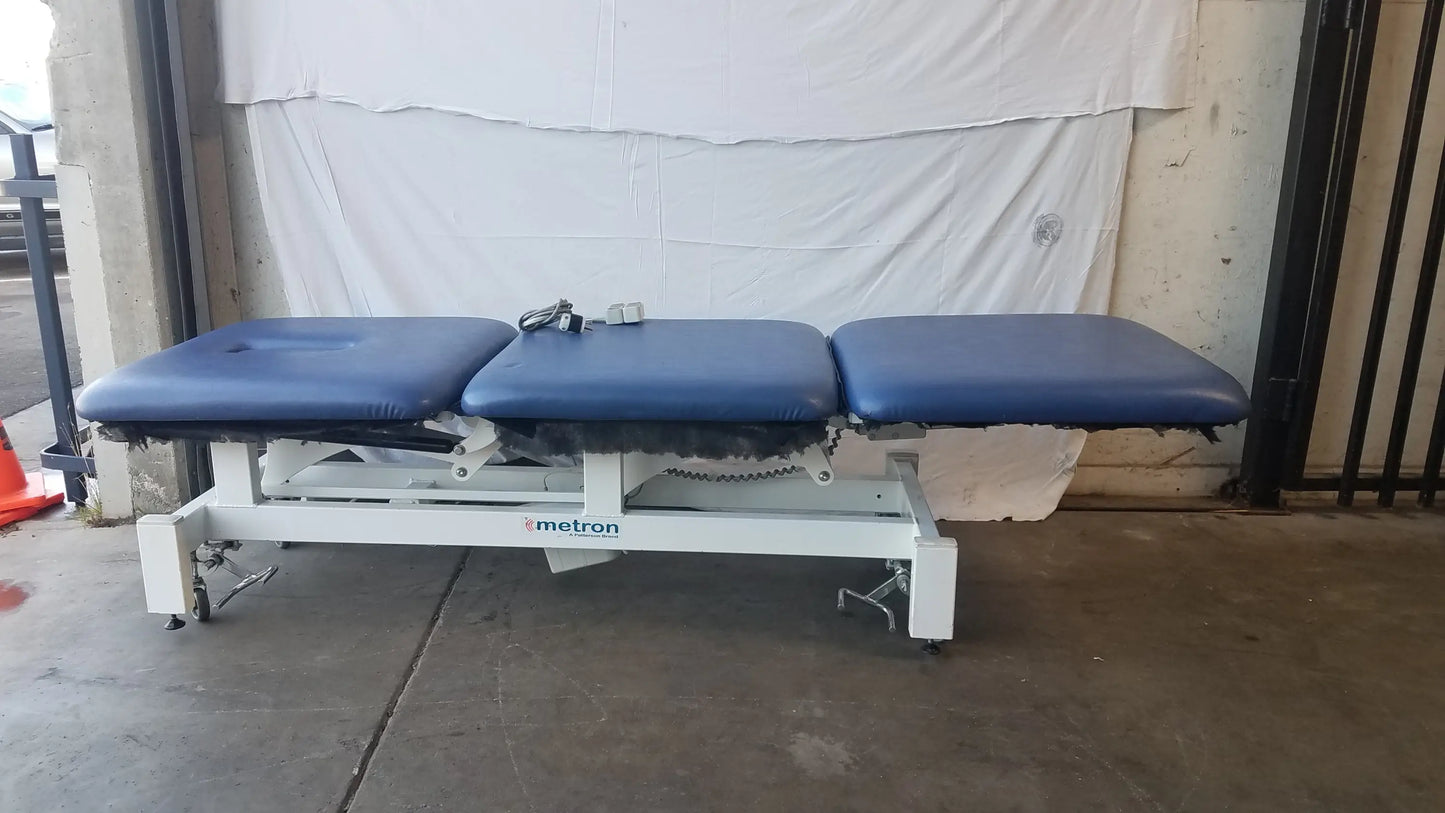 Blue padded three-section electric treatment table with white metal frame and metron branding for medical use