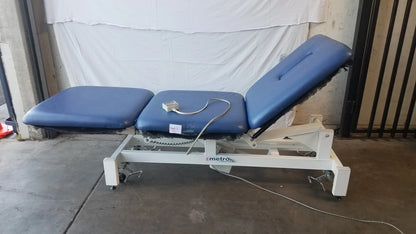 Blue padded three-section metro electric treatment table with white metal frame and casters for adjustable treatment