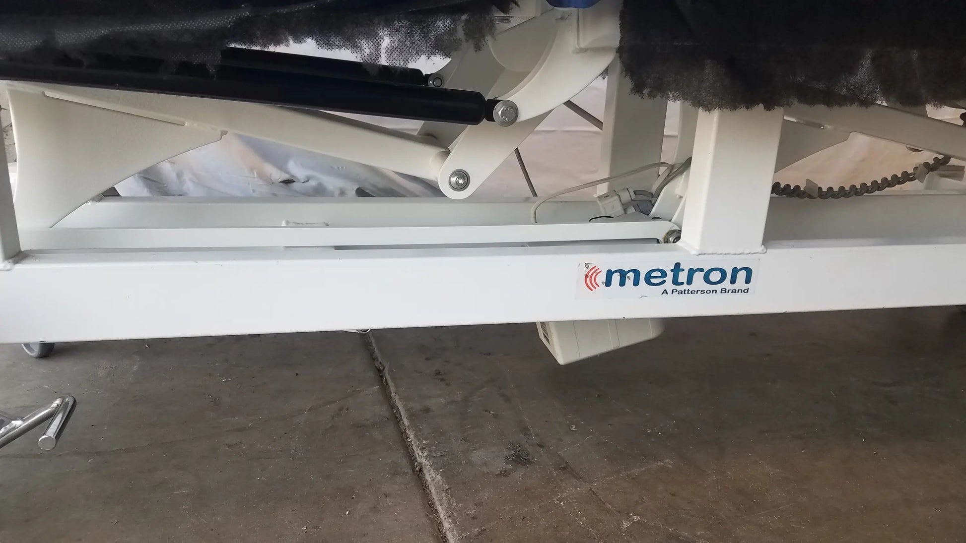 White metal frame of metron 3-section electric treatment table with black hydraulic lift and chain drive system visible from back side