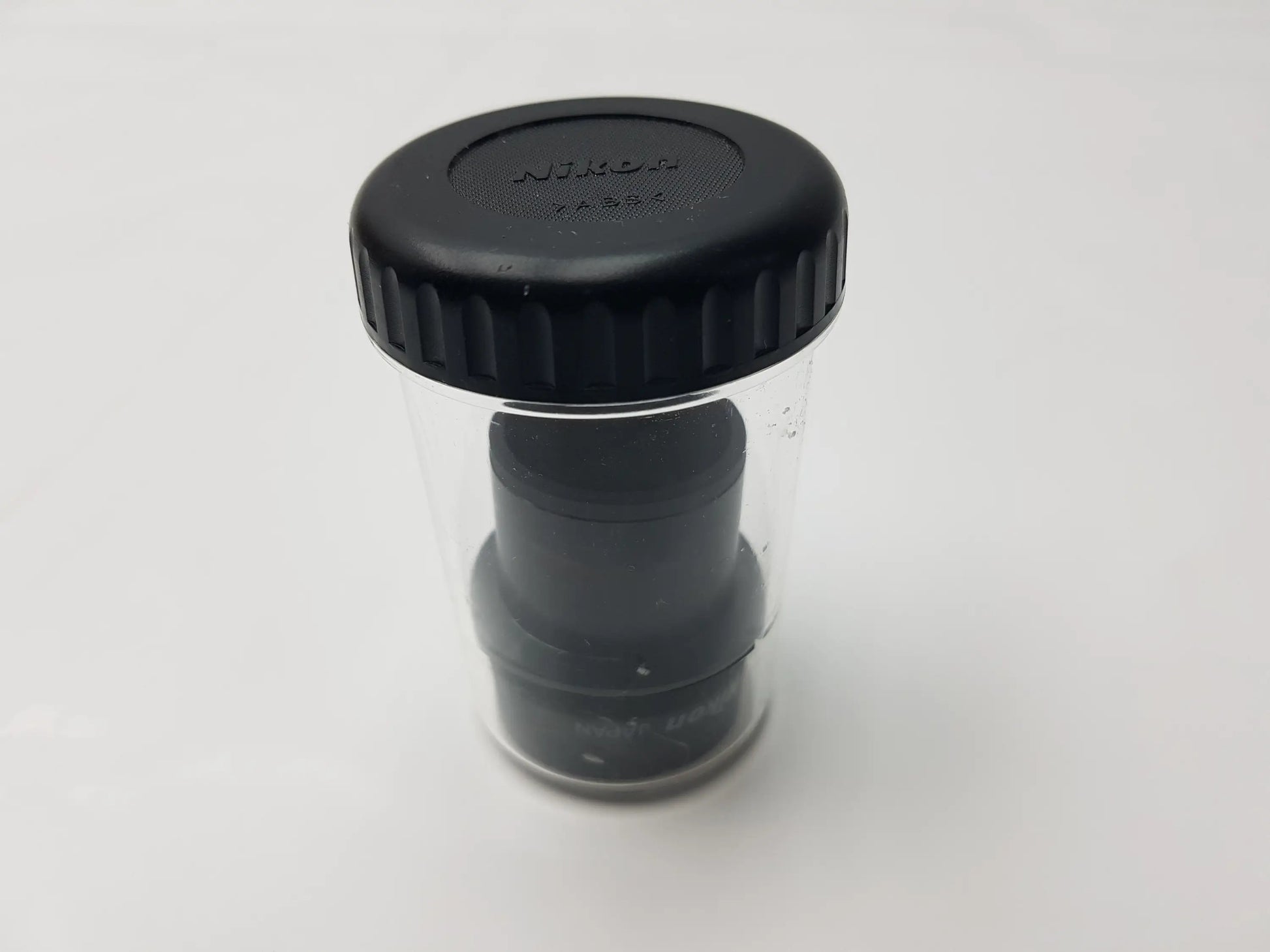 Black Nikon lens cap with textured ridges and embossed branding in clear storage case for compatible Nikon microscope systems