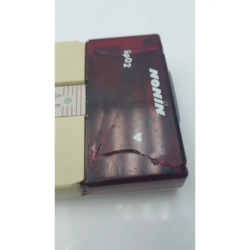 A worn translucent red plastic nikon s902 camera body with scratches and a heart sticker shown with nonin spo2 pulse oximeter model 8500m