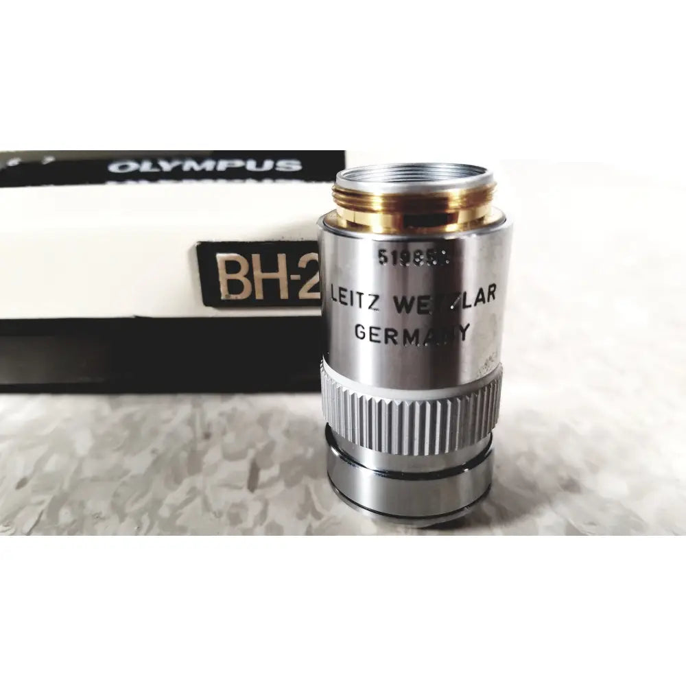 A silver metallic microscope objective lens with gold threaded top beside its olympus bh-2 binocular microscope box
