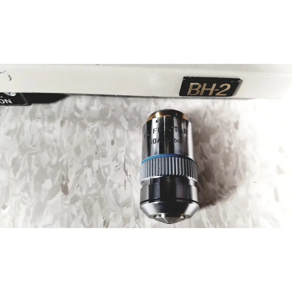 Metallic microscope objective lens with blue ring labeled pl fluor x10 mounted on white olympus bh-2 binocular microscope stage