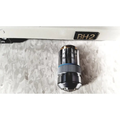 Metallic microscope objective lens with blue ring labeled pl fluor x10 mounted on white olympus bh-2 binocular microscope stage