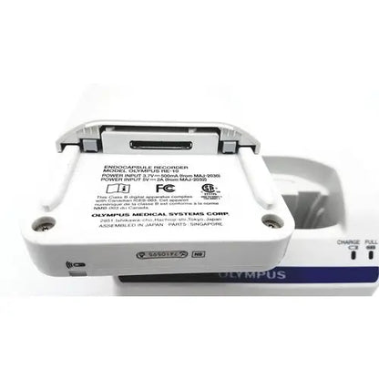 White Olympus capsule recorder digital voice recorder with gray plastic casing and blue MAJ 2032 charging cradle