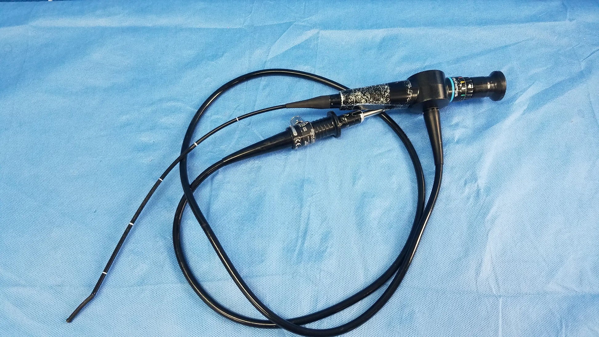 Black flexible fiber optic endoscope with metallic joint and control knobs coiled on blue surgical drape Olympus Rhino-Laryngoscope fiberscope
