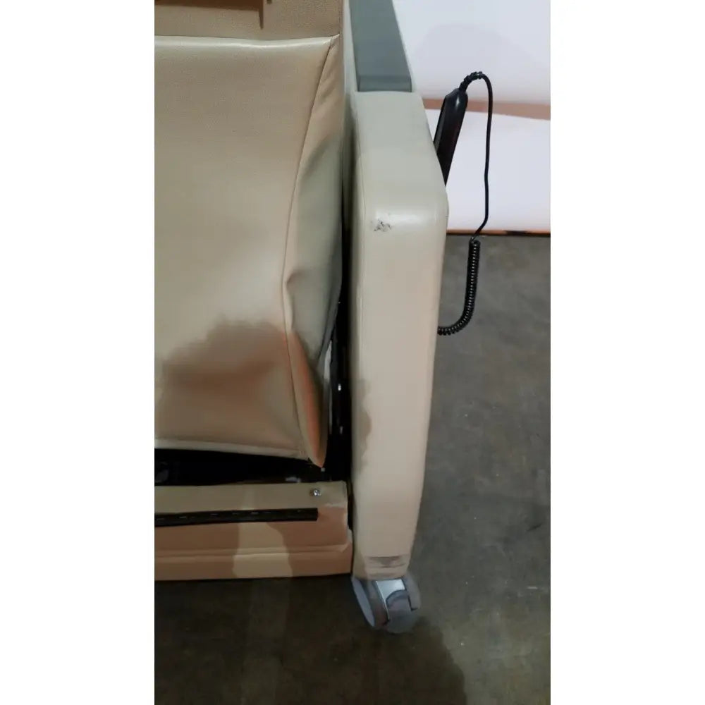 Beige upholstered power recovery recliner with hand control black coiled cable and gray caster wheels for medical use