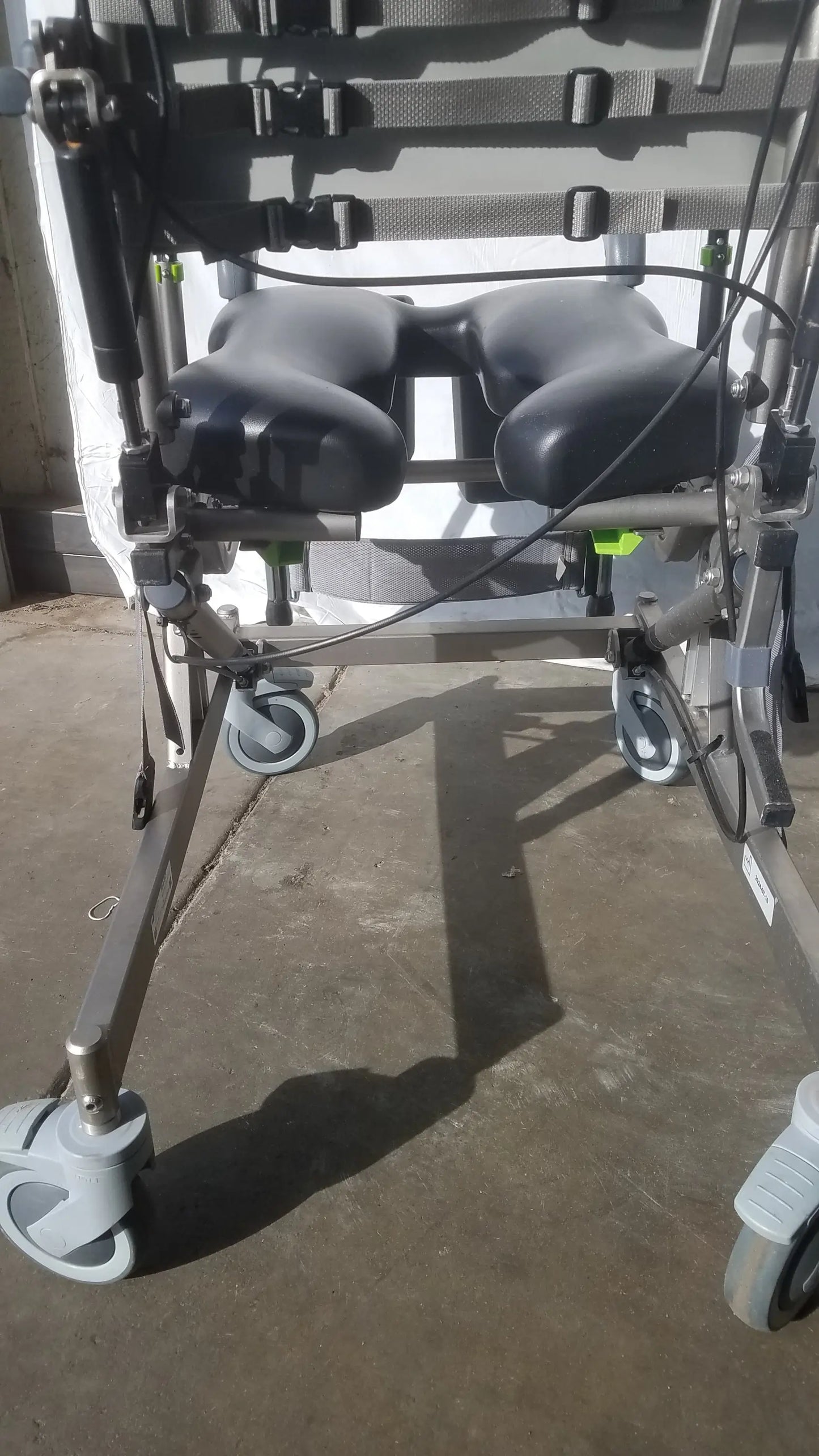 Black padded seat with adjustable straps and gray metal frame on Raz Attendant tilt shower commode chair back side