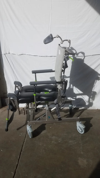 Raz attendant tilt shower commode chair with black padded seat, silver metal frame, adjustable head support, and caster wheels