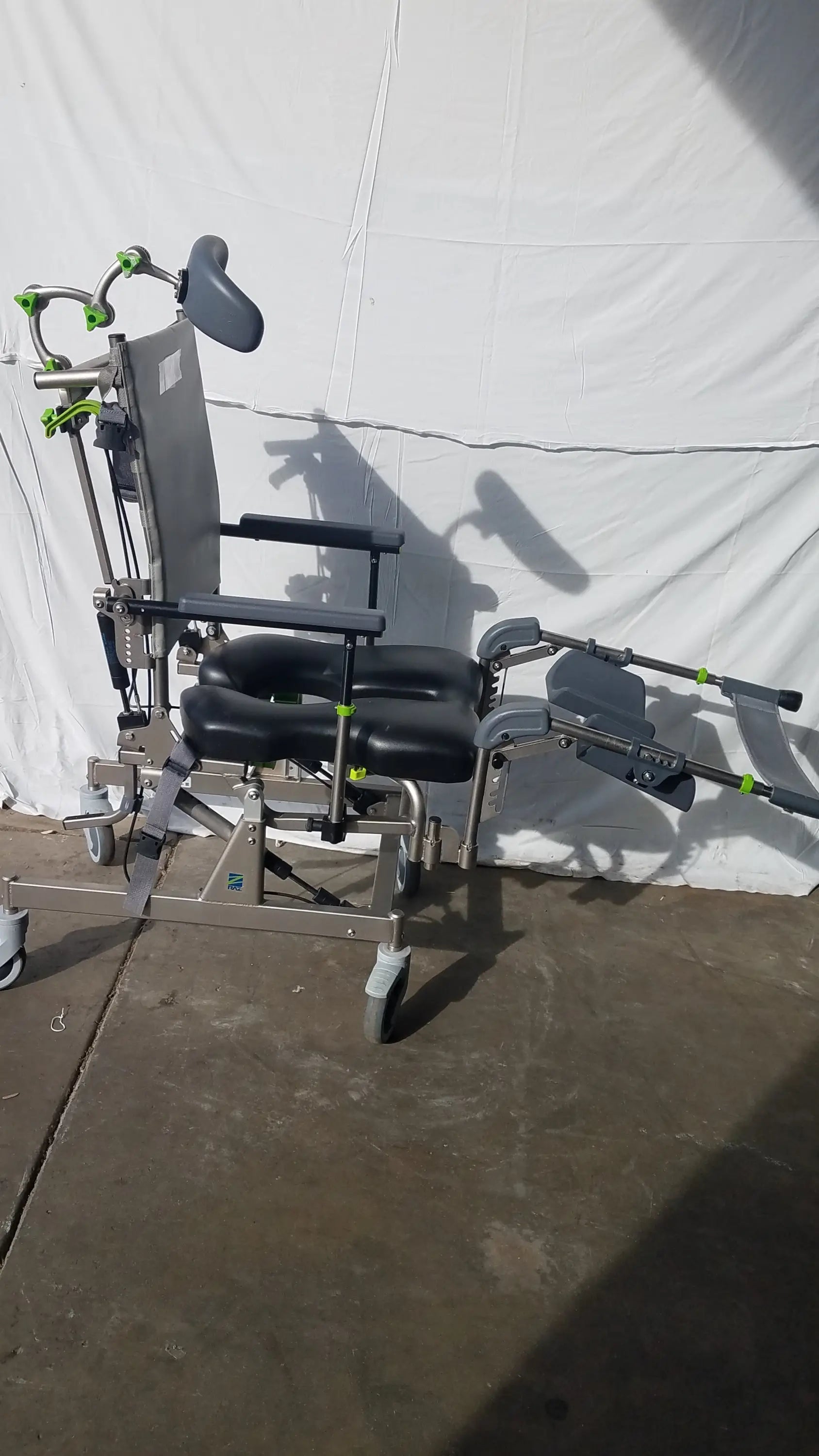 Raz attendant tilt shower commode chair with silver metal frame, black padded cushions, green accents, adjustable headrest and leg supports