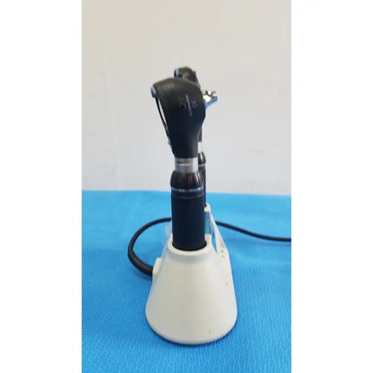 Black and silver medical otoscope with textured grip resting on white riester ri-charger l charging base stand
