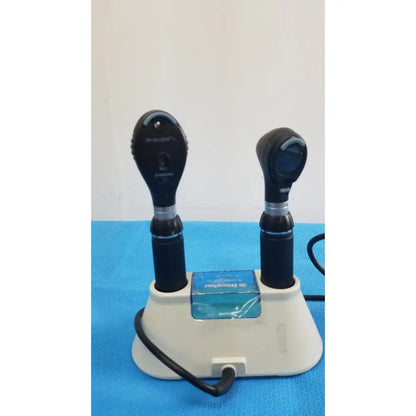 Two black otoscopes with silver accents sit upright in a white riester ri-charger l charging base for reuse medical use
