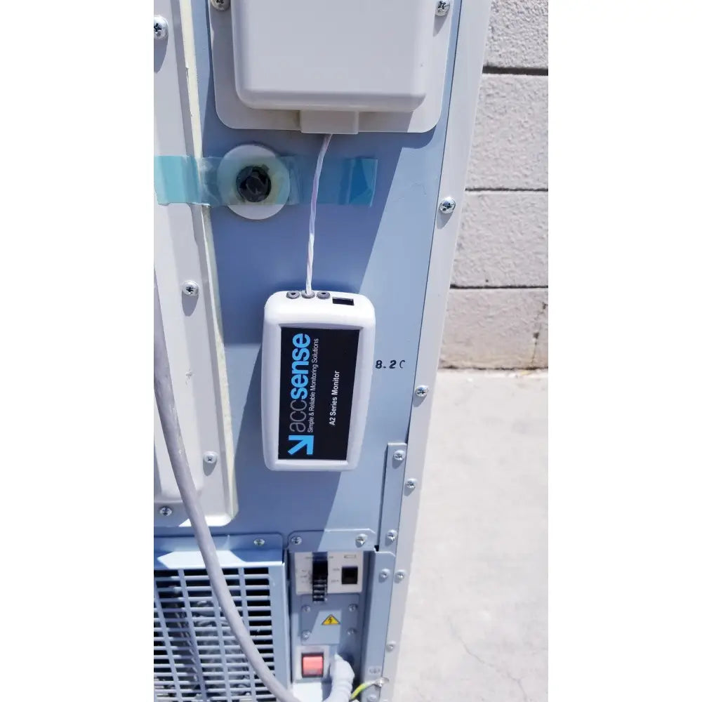 White rectangular Accusense A1 series monitor with black label and blue logo attached to gray Sanyo VIP Series ultra low freezer