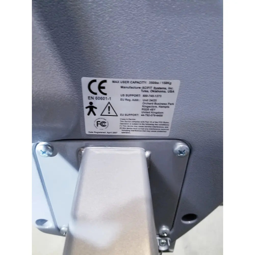 Gray metal safety latch with white label showing manufacturer and certification on scifit sx7000 elliptical commercial lower body trainer