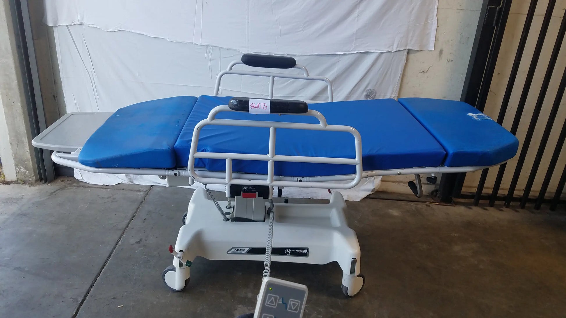 Blue padded TransMotion Medical TMM4 multi purpose stretcher chair with white metal frame and wheels for medical use