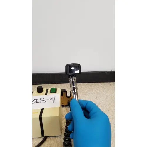 A gloved hand holding a black and silver welch allyn transformer otoscope ophthalmoscope with a coiled cable