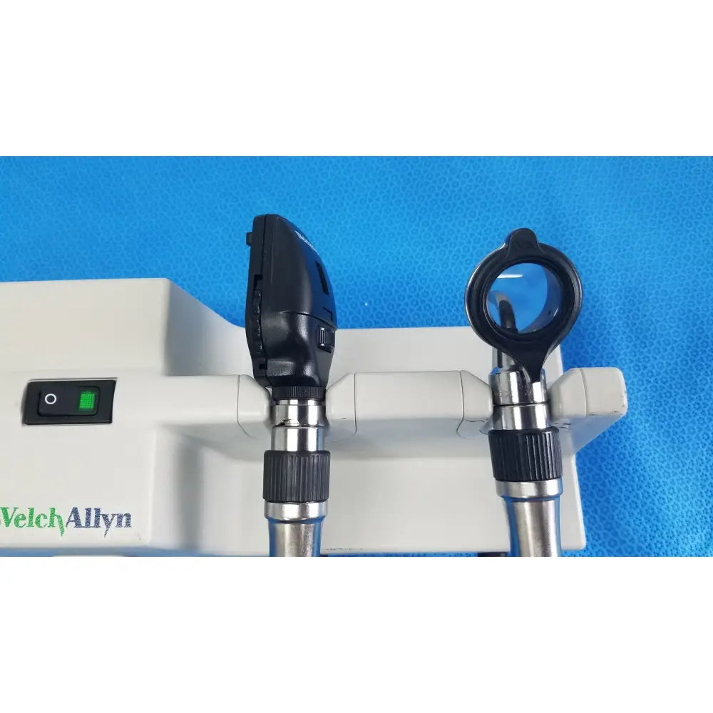 Black and silver welch allyn otoscope with circular lens and angled head mounted on white wall transformer base unit for reuse medical use